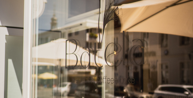 Außenansicht des Cafés Batela. Auf dem Fenster steht Batela und die Straße spiegelt sich.