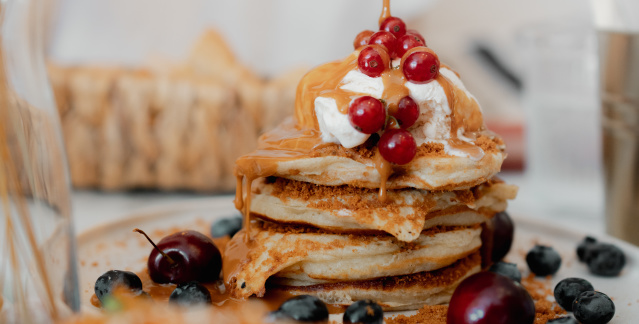 Eine Hand schüttet Sirup aus einem Gläschen auf Pancakes.