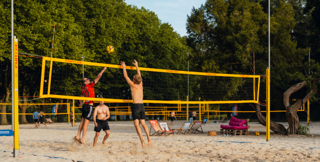 Drei Personen spielen Beachvolleyball im OneBeach. Im Vordergrund stehen zwei Liegestühle.