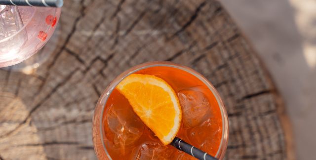 Von oben fotografiert: Ein Aperol Spritz steht mittig und ein Wildberry Lillet links oben auf einem Baumstumpf, welcher auf Sand steht.