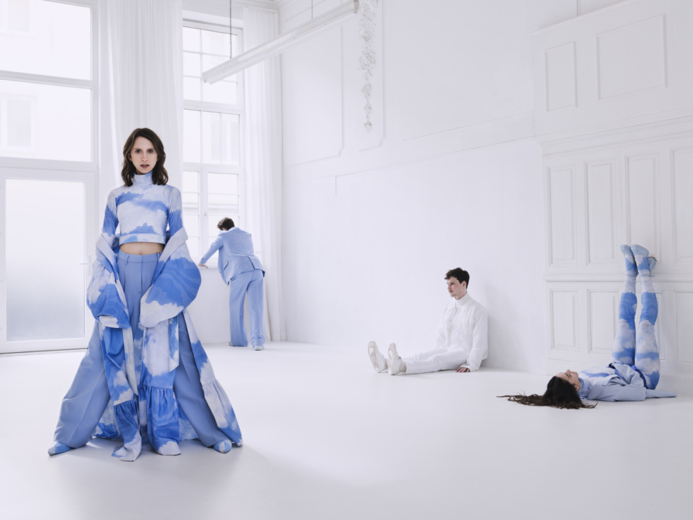 Karoline Weidt Quartett Eine Frau im Vordergrund stehend, ein Mann im Hintergrund angelehnt stehend an eine Fensterbank, ein Mann im Hintergrund auf dem Boden sitzend und eine Frau im Hintergrund auf dem Rücken liegend mit den Beinen an der Wand in einem leerstehenden, weißen R